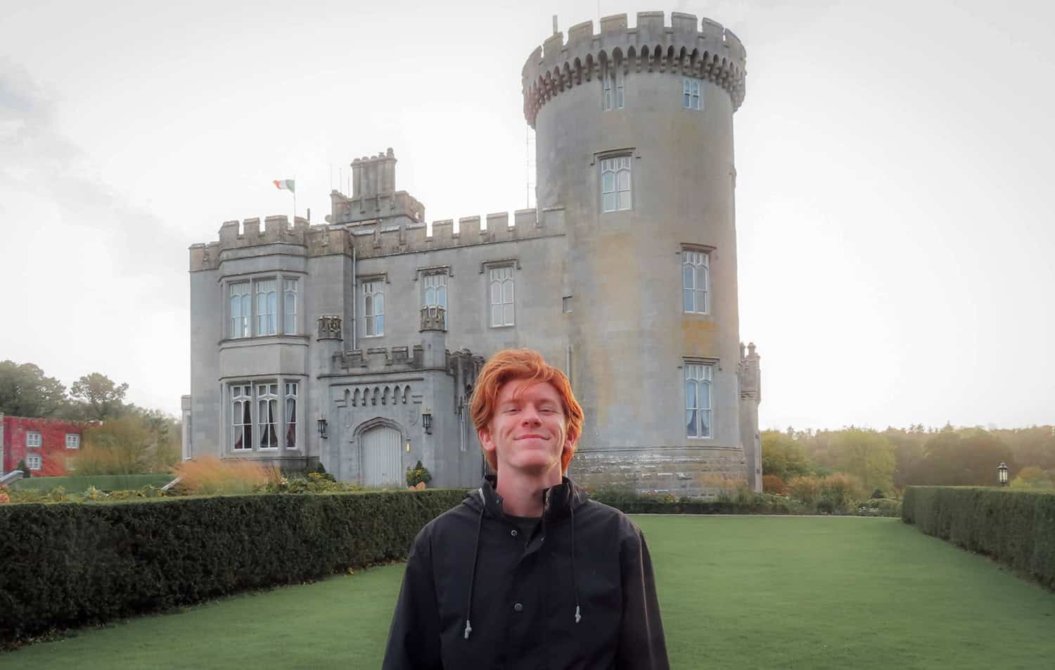 Young man smiling in front of a historic castle with turrets and lush grounds.