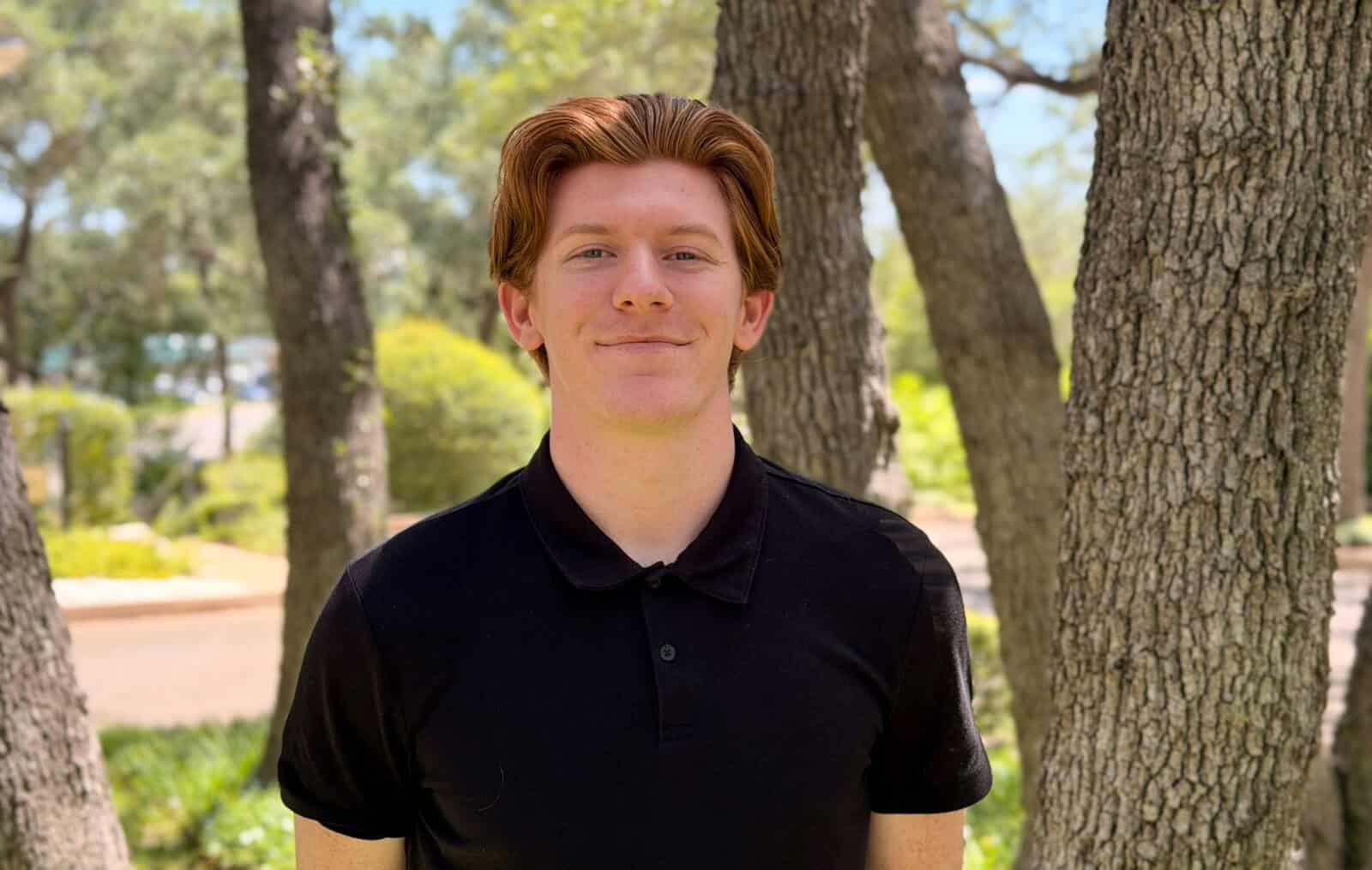 Young man outdoors among trees, smiling, in a black polo shirt.